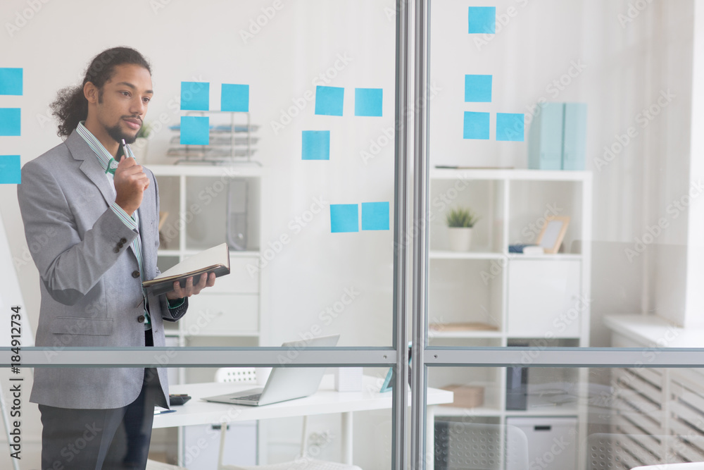Pensive businessman with open notebook standing by transparent noteboard in office Pensive businessman with open notebook standing by transparent noteboard in office