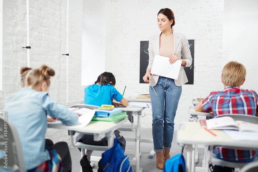 Fototapeta premium Strict teacher walking down between desks while schoolkids carrying out individual task