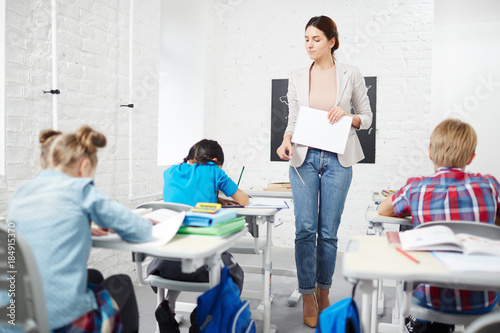 Strict teacher walking down between desks while schoolkids carrying out individual task