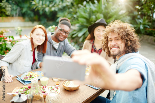 Young happy friends sitting by served table and making selfie during dinner in garden room