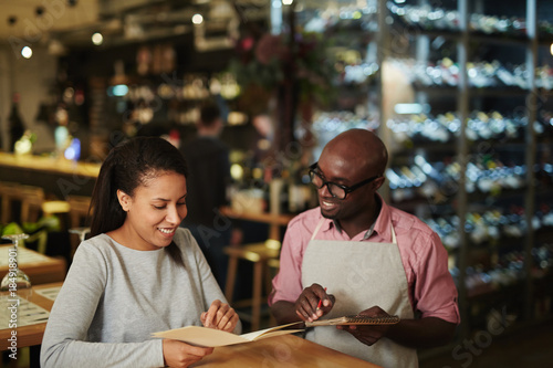 Young waiter writing down order of visitor and consulting her about new food