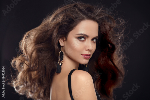 Fashion portrait of gorgeous sexy brunette woman with long healthy hair style and evening makeup, black earrings jewelry looking at camera isolated on black studio background with back light.