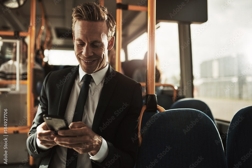 Smiling young businessman standing on a bus sending text messsages - obrazy, fototapety, plakaty 