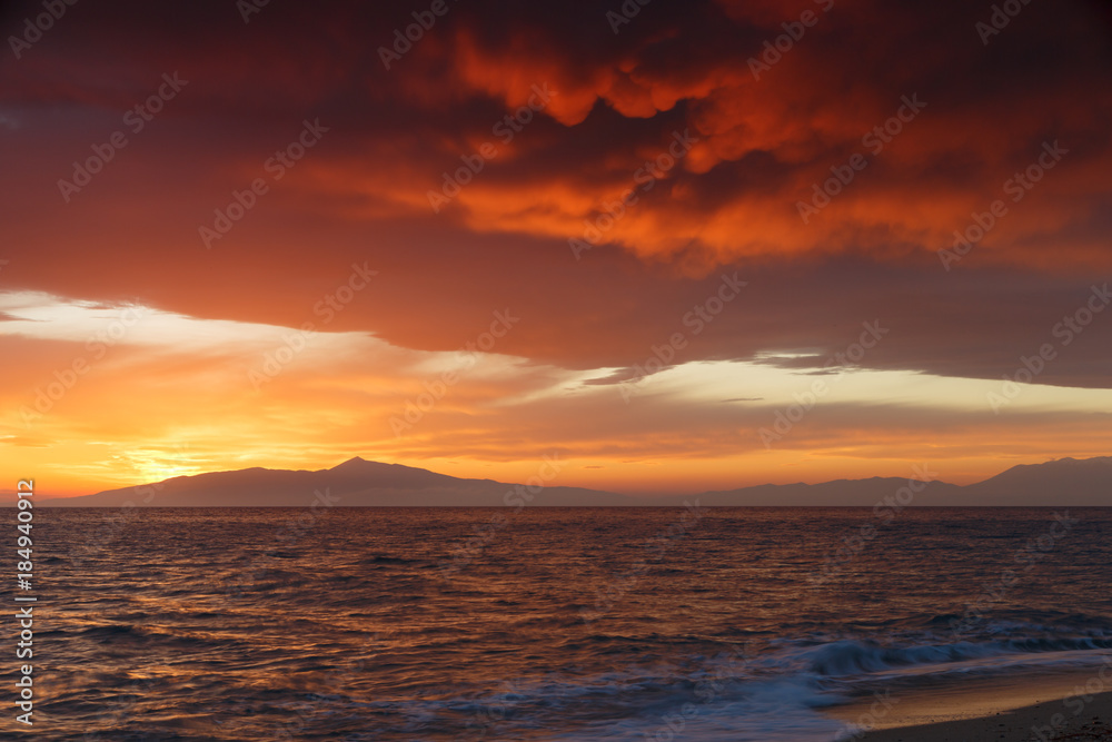 Crimson sunset on the shore of the orange sky and calm sea witt view at the mountains