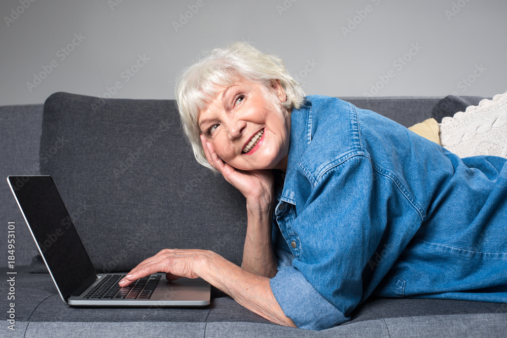 Portrait of old curious lady resting on couch with laptop. She is leaning head on hand while fingers are typing on computer Portrait of old curious lady resting on couch with laptop. She is leaning head on hand while fingers are typing on computer