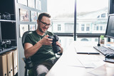 Portrait of cheerful unshaven man looking at camera while holding it in hands. He locating at desk in room. Job concept