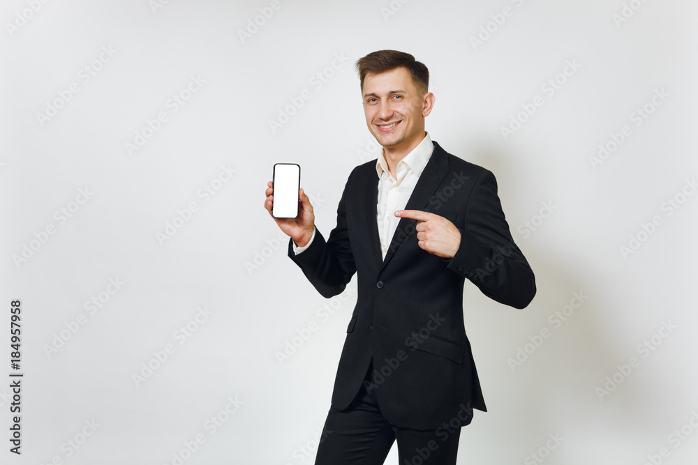 Young successful handsome rich business man in black suit pointing at modern mobile phone isolated on white background for advertising. Concept of money, achievement, career and wealth in 25-30 years. Young successful handsome rich business man in black suit pointing at modern mobile phone isolated on white background for advertising. Concept of money, achievement, career and wealth in 25-30 years.