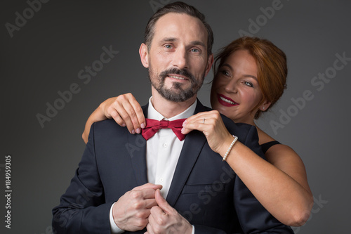 Portrait of caring middle-aged woman is adjusting bowtie on male neck. She is embracing man in suit from behind and smiling. Preparation for anniversary party concept. Isolated