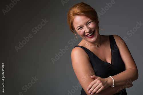Waist up portrait of happy middle-aged woman standing in elegant black dress. She is looking at camera and laughing. Isolated and copy space
