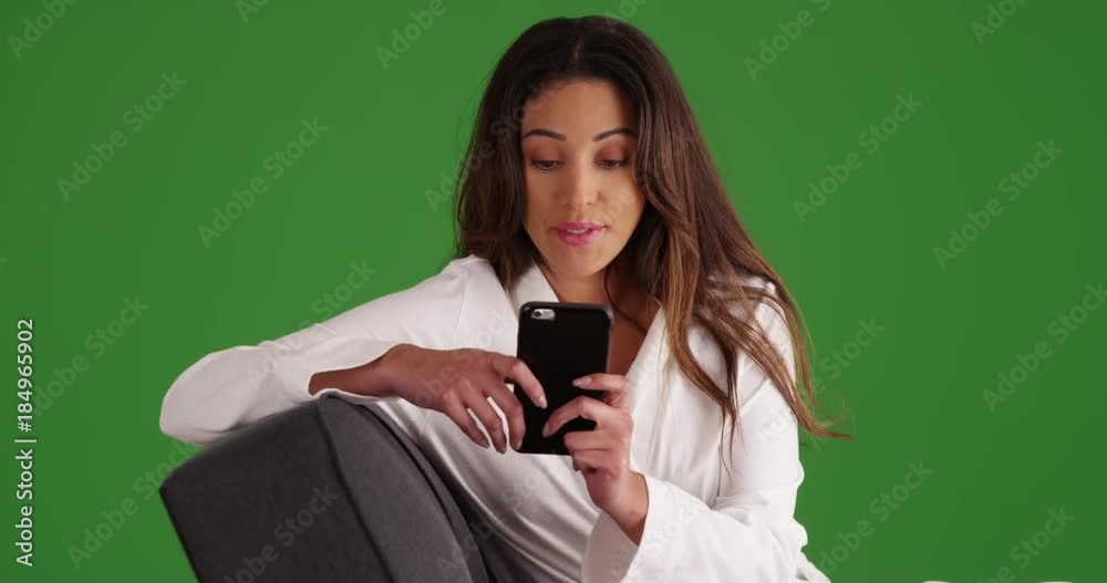 Attractive Latina female in white bathrobe lounging on sofa, texting someone with smart phone on green screen. On green screen to be keyed or composited. Attractive Latina female in white bathrobe lounging on sofa, texting someone with smart phone on green screen. On green screen to be keyed or composited.