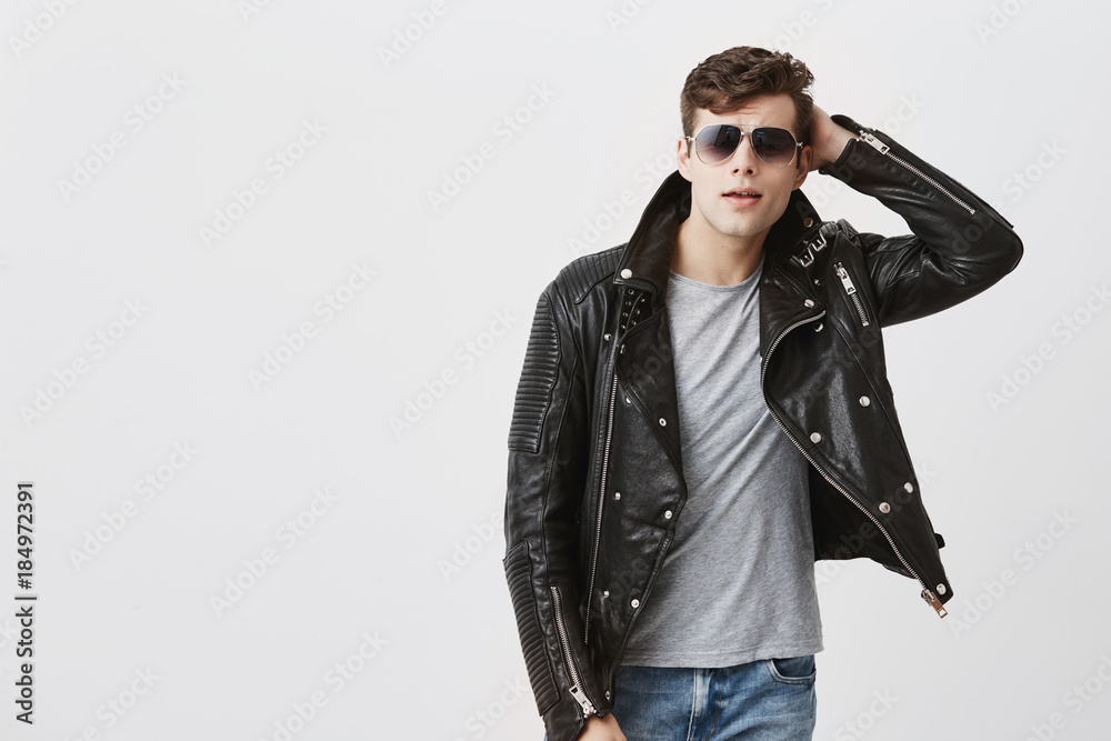 Horizontal portrait of attractive Caucasian man with sunglasses on, stylish haircut, dressed in black leather jacket, looks seriously into camera. Muscular handsome male model poses in studio Horizontal portrait of attractive Caucasian man with sunglasses on, stylish haircut, dressed in black leather jacket, looks seriously into camera. Muscular handsome male model poses in studio