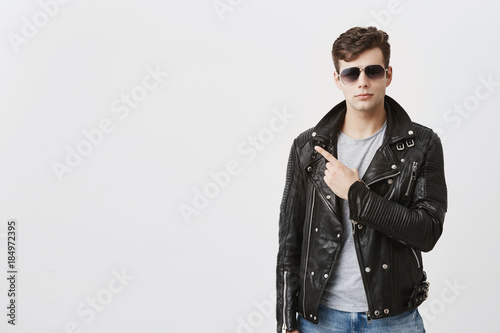 Confident good-looking guy in black leather jacket with sunglasses on, indicates with fore finger at copy space for advertisment or promotional text. Handsome stylish male points into distance