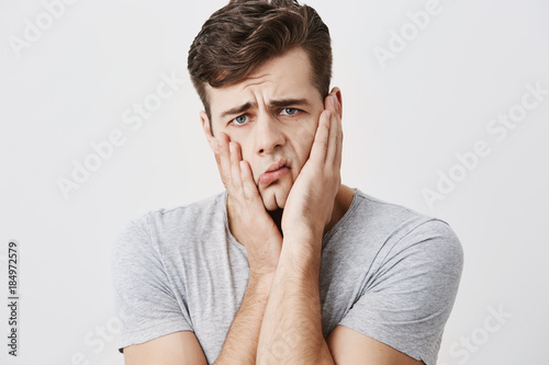 Shocked stunned emotional man keeps hands on cheeks, being troubled to listen advices of his parents isolated against blank studio wall background. European male can`t believe in shocking news