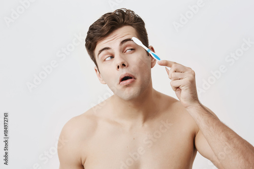 Positive naked muscular guy with trendy hairstyle, healthy skin, making faces, concentrated on combing his eyebrows with toothbrush. Attractive good-looking male posing against gray background.