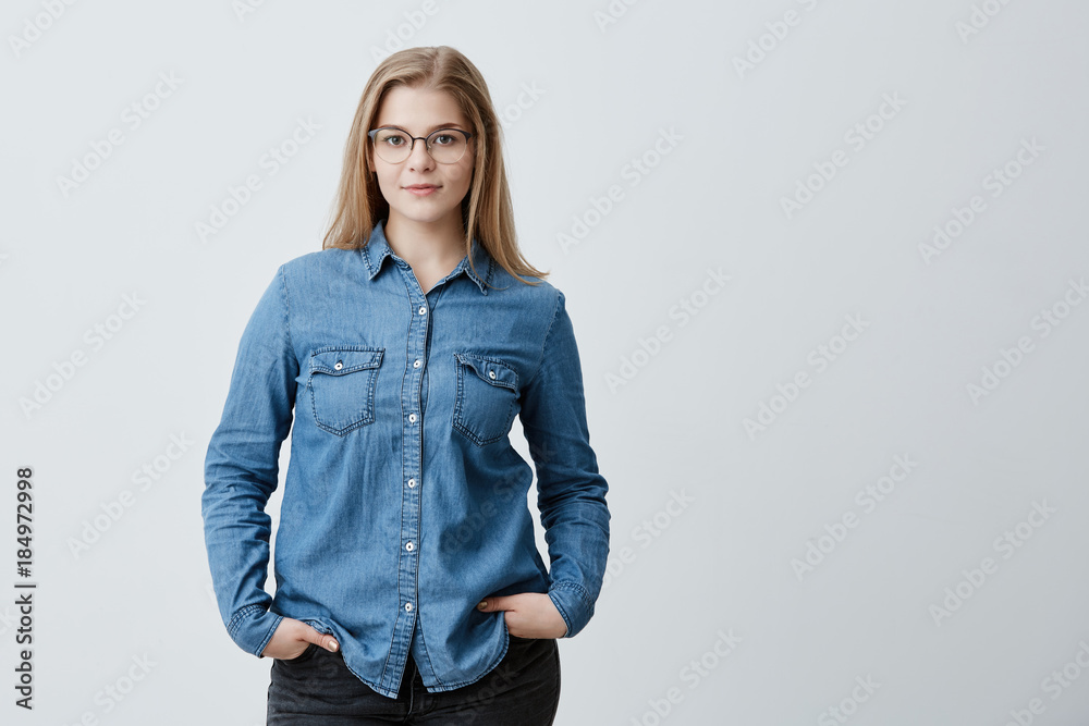 Stylish pretty female student with straight blonde hair and spectacles dressed in denim shirt posing indoors, holding her hands in pockets. People, beauty and lifestyle concept