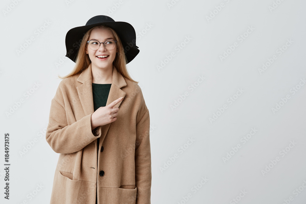 Fototapeta premium Beautiful smiling woman in coat over green sweater and eyeglasses pointing at blank white wall while demonstrating something. Cheerful young female indicating with fore finger on gray background.