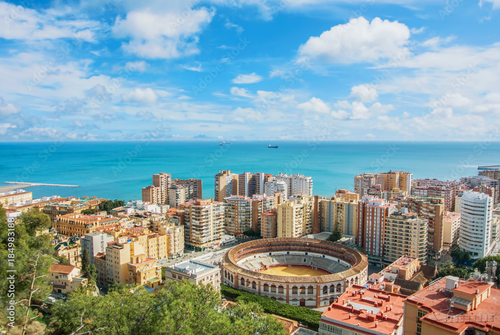 Beautiful iconic famous panoramic view of Malaga city center, a bullring, hotels, houses and Mediterrain sea seascape with cargo ships on sunny cloudy evening from Gibralfaro Castle, Andalusia, Spain.