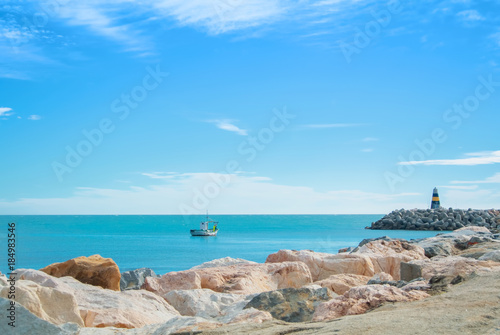 Panoramic seascape of Mediterranean sea with a lonely fishing boat and a small black and yellow lighthouse at Benalmadena port, Malaga province, Spain. Big stones at sandy bay beach at foreground.
