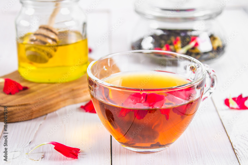 Fototapeta premium A cup of tea, in the background of a bank of honey and a jar with a black herbal floral tea on a white wooden table.