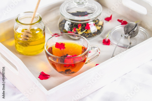 A cup of tea, a can of honey and a jar of black herbal tea on a white tray in bed.