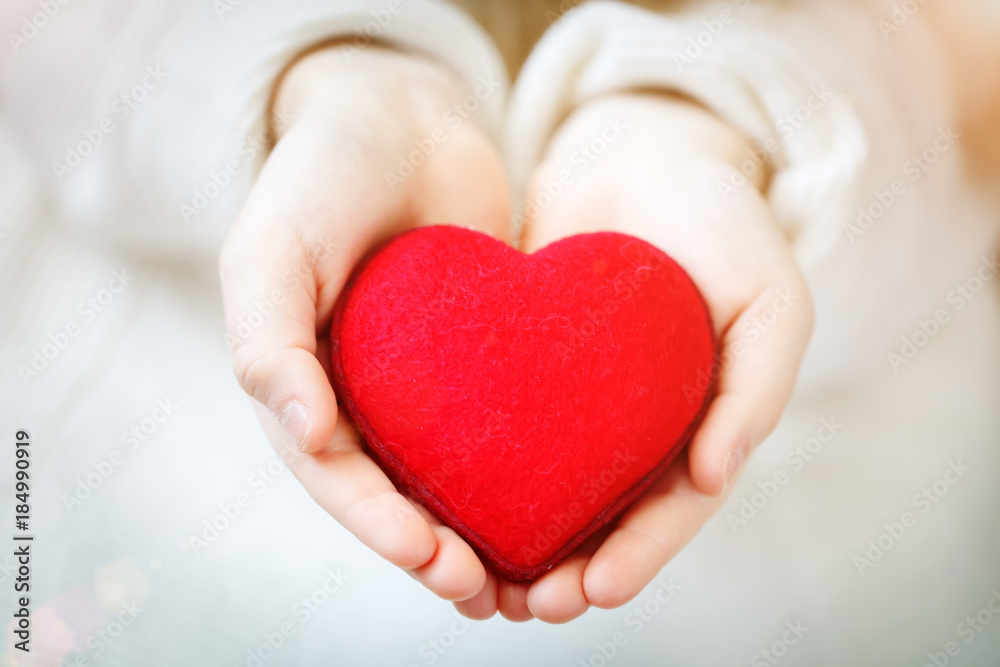 Red heart in hands of the little girl. Symbol of love and family.Valentines day card. Mother's day. Backgrounds for social posters. Selective focus. Red heart in hands of the little girl. Symbol of love and family.Valentines day card. Mother's day. Backgrounds for social posters. Selective focus.