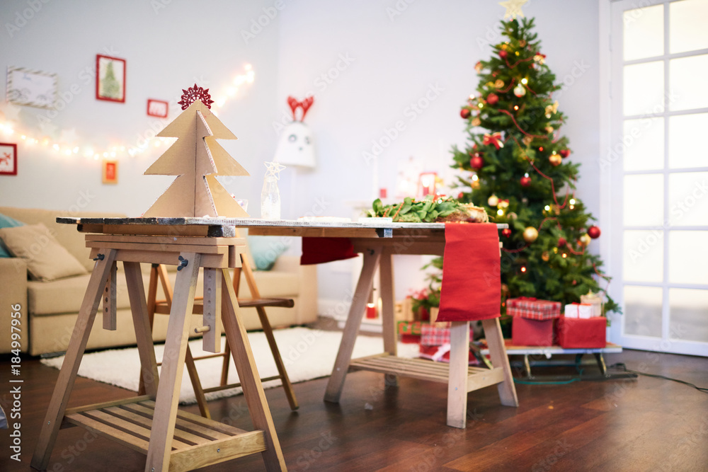 Interior of cozy living room decorated for celebrating Christmas: small handmade pictures hanging on wall, desk with unfinished decorations, bushy Christmas tree Interior of cozy living room decorated for celebrating Christmas: small handmade pictures hanging on wall, desk with unfinished decorations, bushy Christmas tree