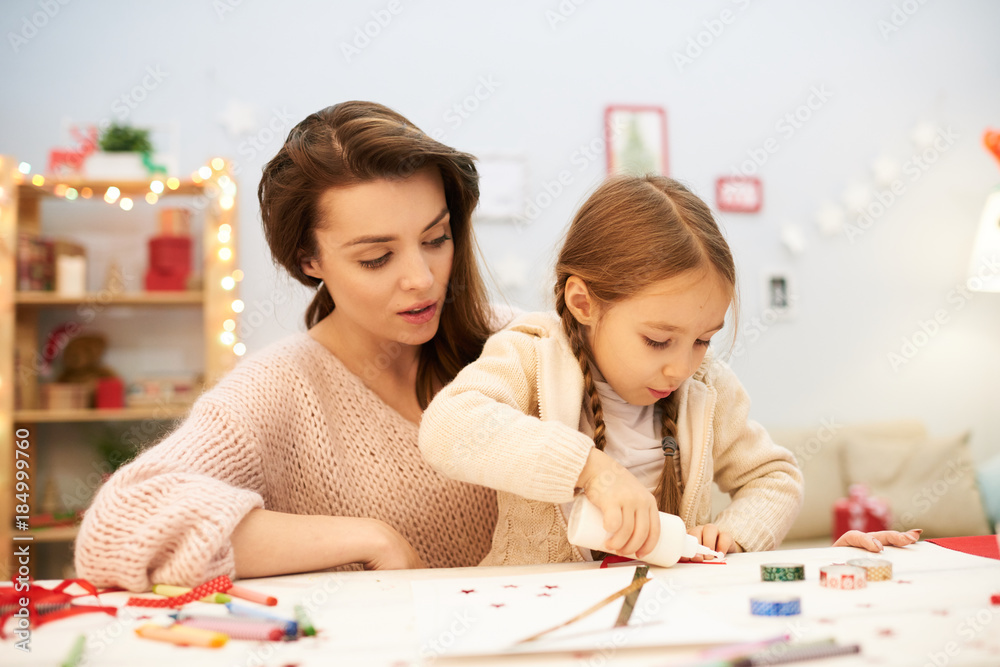 Fototapeta premium Pretty little girl sitting on laps of her mother and using glue in order to make Christmas card, interior of cozy living room on background