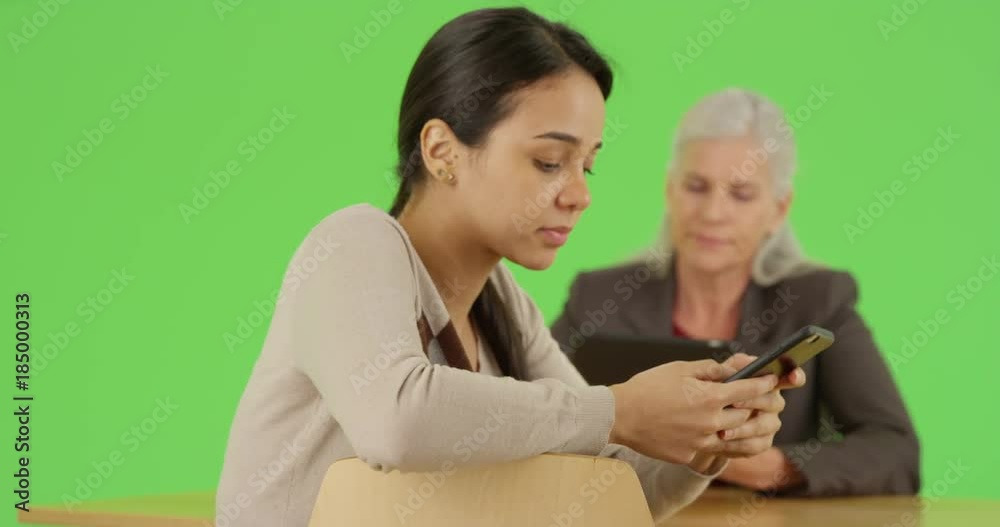 A young Latina businesswoman responds to emails on her smart phone while her boss uses her tablet on green screen. On green screen to be keyed or composited. 