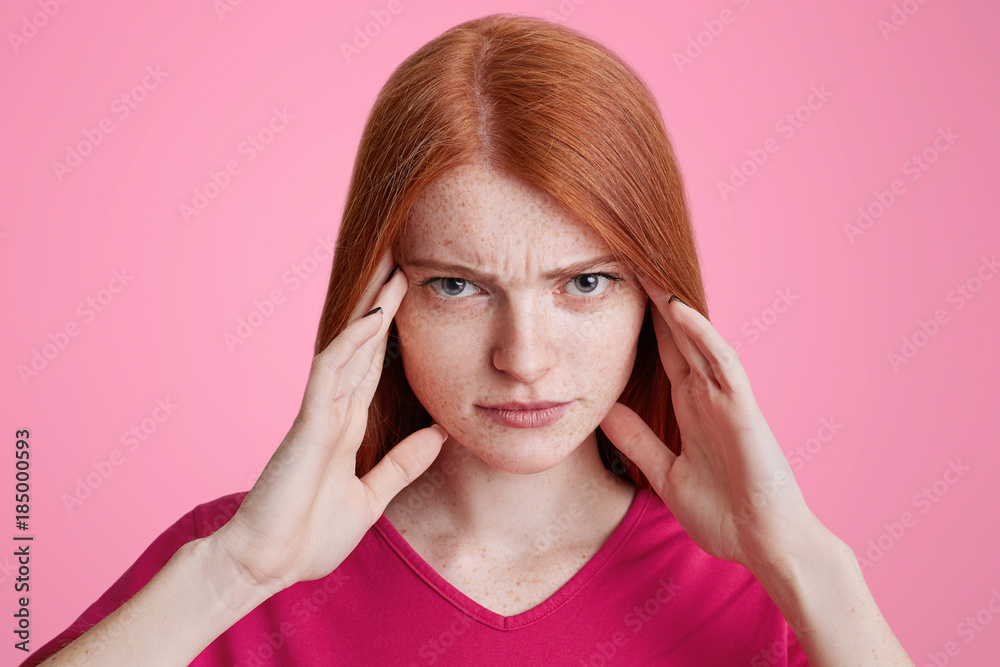 Portrait of red haired freckled female tries to concentrate, has serious expression, keeps hands on temples, has headache, wears bright pink sweater, poses in studio. Facial expressions concept