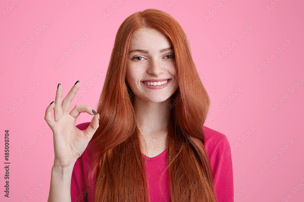 Obraz premium Photo of positive ginger female with long hair, shows okay symbol over pink background, expresses her approval, being in good mood, satisfied with something. People and body language concept