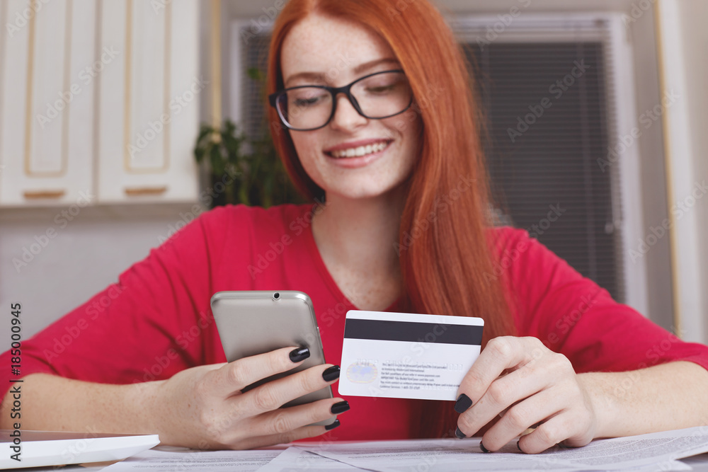 Redhaired pretty female model in eyewear holds smart phone and credit card, makes online purchase or shopping in internet store, has happy expression. Online payment. Mobile payments concept Redhaired pretty female model in eyewear holds smart phone and credit card, makes online purchase or shopping in internet store, has happy expression. Online payment. Mobile payments concept