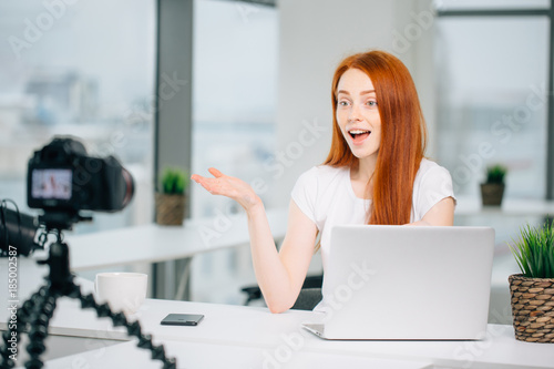 Young woman recording video on camera mounted on tripod for her vlog. Pretty woman smiling at camera sitting at table with laptop photo