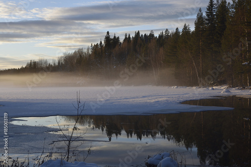 Midwinter day in the North of Sweden with fog over a frozen lake with a open spot of calm water reflecting the forest and sky.