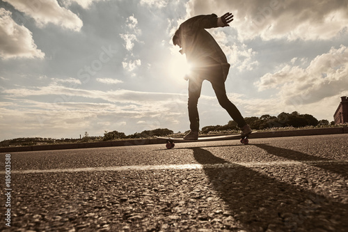 Back outdoor view of unrecognizable hipster guy sliding on skateboard along textured copy space asphalt. Skateboarder riding longboard on empty road in the morning. Flare sun, warm toned image photo