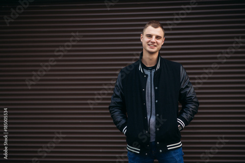 A young guy in a black jacket against a background of dark striped walls posing and smiling at the photographer