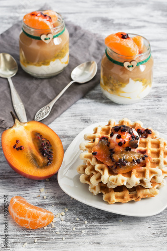 Homemade waffles are poured tamarillo on a rustic background. Yogurt with passion fruit and apple puree in small jars.