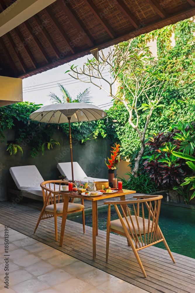 Beautiful cozy luxury breakfast for two at the private pool, Bali,Indonesia.A wooden table with an abundant healthy breakfast on the background of the pool and tropical plants.