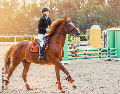 Young girl in uniform jumping with sorrel horse. Blond pretty little girl going jump a hurdle in a competition. Girl with red horse during equestrian showjumping.