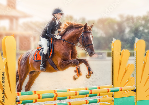 Young girl in uniform jumping with sorrel horse. Blond pretty little girl going jump a hurdle in a competition. Girl with red horse during equestrian showjumping.