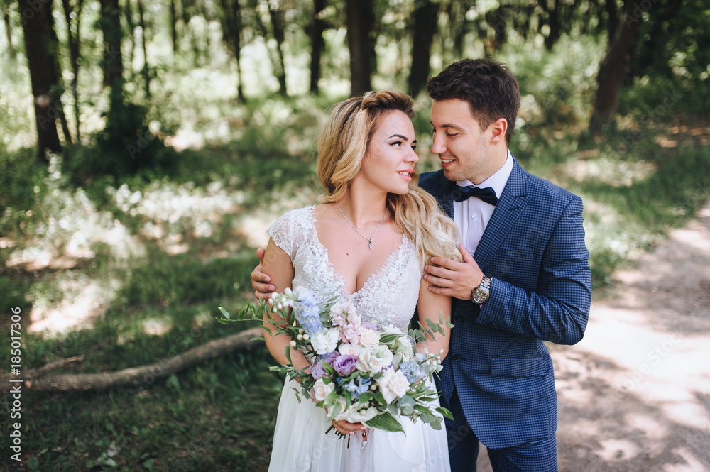 The groom tenderly embraces the bride by the shoulders. The bride is holding a bouquet. Wedding walk in the forest. Lovers look at each other. The groom tenderly embraces the bride by the shoulders. The bride is holding a bouquet. Wedding walk in the forest. Lovers look at each other.