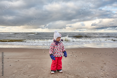 Little girl wear jacket and hat walk is playing at the beach near baltic sea on cloudy sky in winter time
