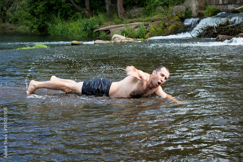 Warm summer day, rest on the nature. A young man is swimming on a very shallow river, he is funny and cheerful.