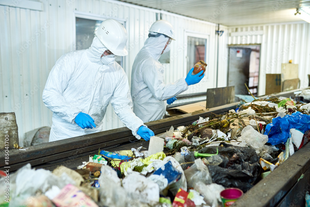 Portrait of two workers wearing biohazard suits working at waste processing plant sorting trash on conveyor belt, copy space Portrait of two workers wearing biohazard suits working at waste processing plant sorting trash on conveyor belt, copy space