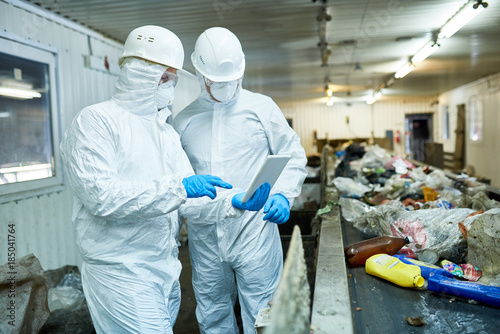 Portrait of two workers wearing biohazard suits using digital tablet standing by conveyor belt at waste processing plant , copy space