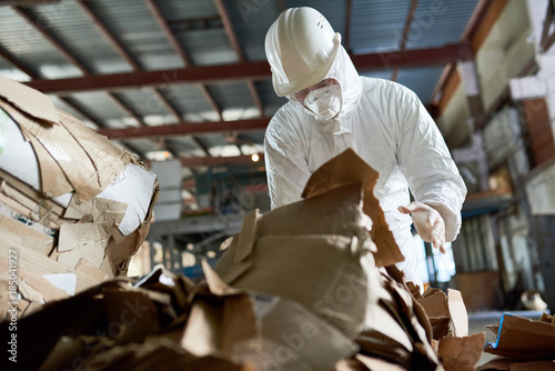 Portrait of factory worker wearing biohazard suit sorting reusable cardboard on recycling plant, copy space