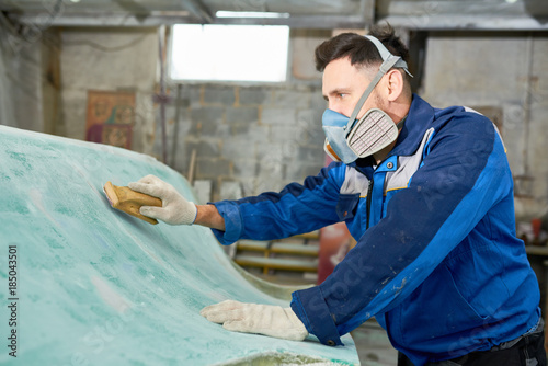 Side view portrait of young  man wearing respirator repairing boat in yacht workshop using polishing tool, copy space