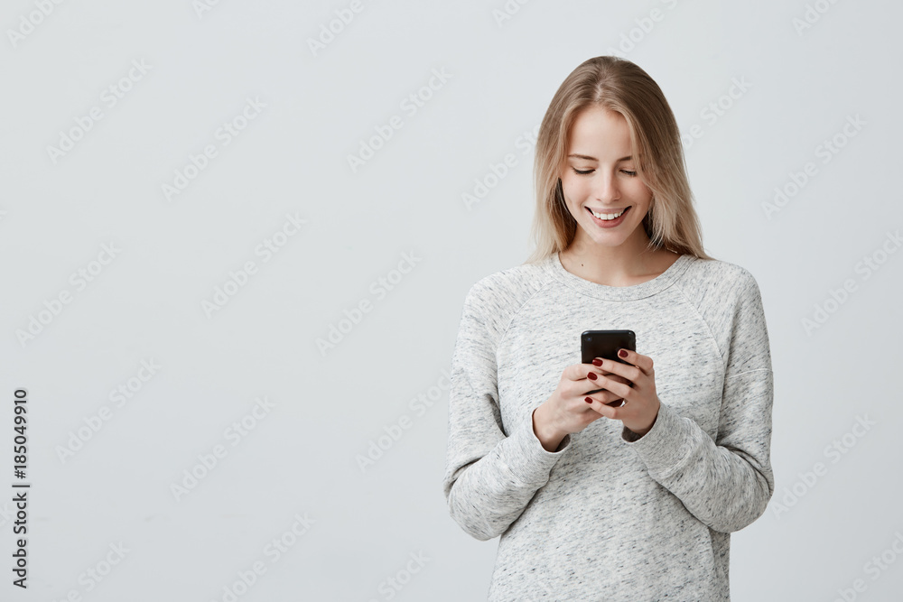Blonde smiling young woman demonstrating white teeth using cell phone, messaging, being happy to text with her boyfriend, looking at screen of smartphone. Modern technologies and communication Blonde smiling young woman demonstrating white teeth using cell phone, messaging, being happy to text with her boyfriend, looking at screen of smartphone. Modern technologies and communication
