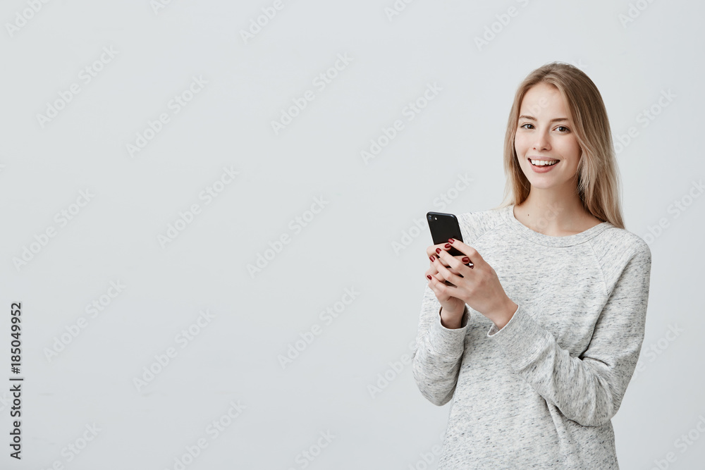 Cheerful young blonde-haired female student with cute smile posing indoors, using cell phone, checking newsfeed on her social network accounts. Pretty girl surfing internet on mobile Cheerful young blonde-haired female student with cute smile posing indoors, using cell phone, checking newsfeed on her social network accounts. Pretty girl surfing internet on mobile