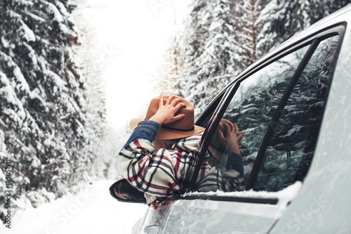 Handsome woman hanging out of the car window, hold vintage hat and enjoying beautiful winter snow road in forest. Traveler in winter nature