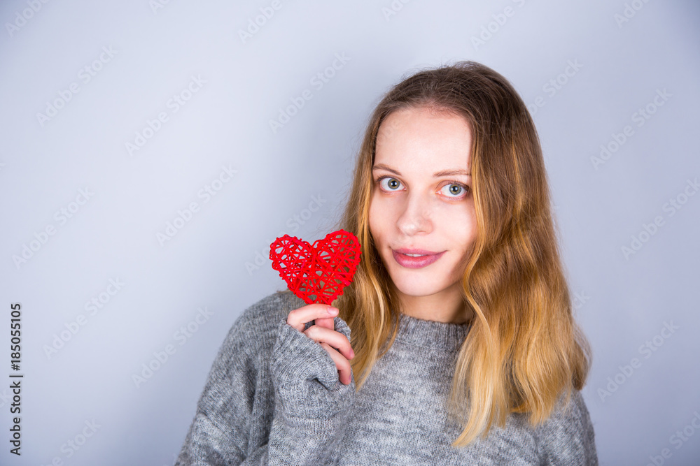 Smiling young blond woman in gray sweater holding red wicker heart on light background Smiling young blond woman in gray sweater holding red wicker heart on light background